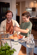 Load image into Gallery viewer, Smiling boy uses tongs to serve spaghetti with red sauce from a blue pan as his dad laughs behind him.