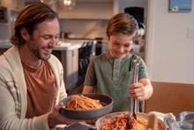 Load image into Gallery viewer, A smiling father serves a bowl of spaghetti to his son who eagerly reaches for more with a pair of tongs.