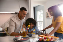 Load image into Gallery viewer, A father and mother smile at their son as he assembles an appetizer skewer in their bright kitchen.