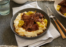 Load image into Gallery viewer, Tender pot roast topped with pepperoncini peppers served over a generous helping of mashed potatoes in a black bowl.