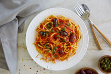 Load image into Gallery viewer, A plate of Pasta Puttanesca made with Kalamata olives and capers and utensils
