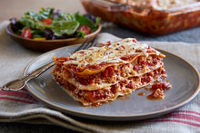 Load image into Gallery viewer, A cheesy slice of lasagna sits on a blue plate, fresh from a dish of lasagna in the background.
