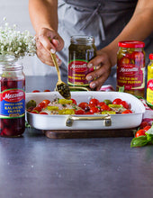 Load image into Gallery viewer, A woman adds capers to a white baking dish filled with seasoned chicken breasts, cherry tomatoes, pepper rings, and olives.