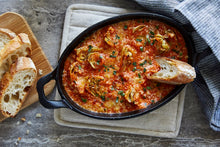 Load image into Gallery viewer, A cast-iron skillet holds cheesy artichoke dip topped with fresh parsley, and a slice of crusty bread rests on the edge. 

