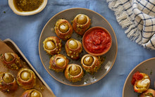 Load image into Gallery viewer, Pesto-stuffed olives wrapped in dough and baked until golden brown are served on a plate with small bowls of pesto and tomato sauce. 

