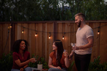 Load image into Gallery viewer, Three friends laugh and enjoy drinks at an outdoor evening gathering illuminated by string lights. 

