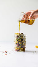 Load image into Gallery viewer, Hand pouring a jar of olive oil into a jar of mixed olives
