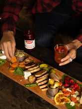 Load image into Gallery viewer, Man assembles a charcuterie board with sliced baguette, cheeses, olives, pickled vegetables, and a cocktail with a brandied cherry. 

